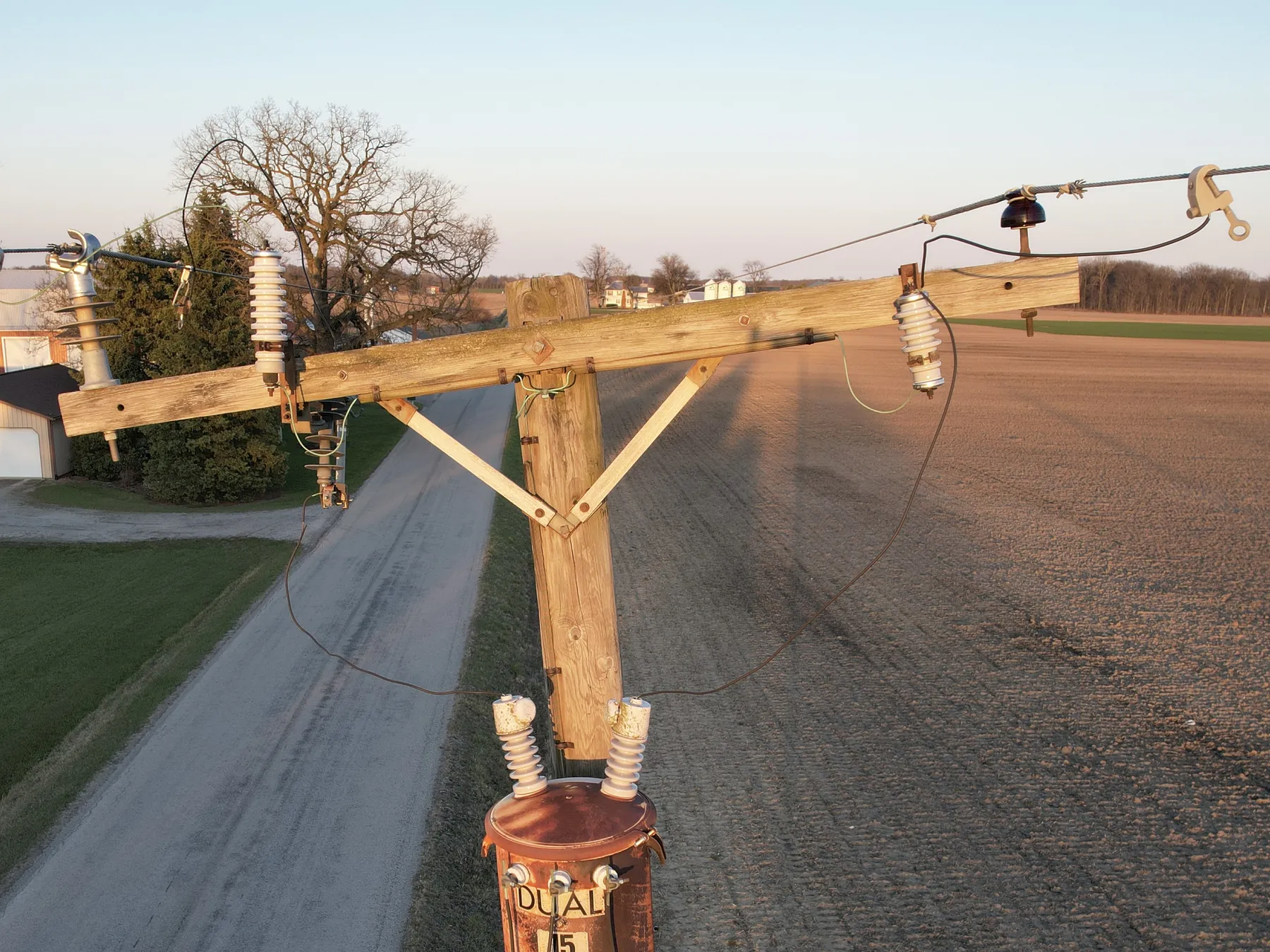 Before: drone view of an old, weathered wooden pole with a worn crossarm and a rusty transformer beside a rural gravel road at sunset