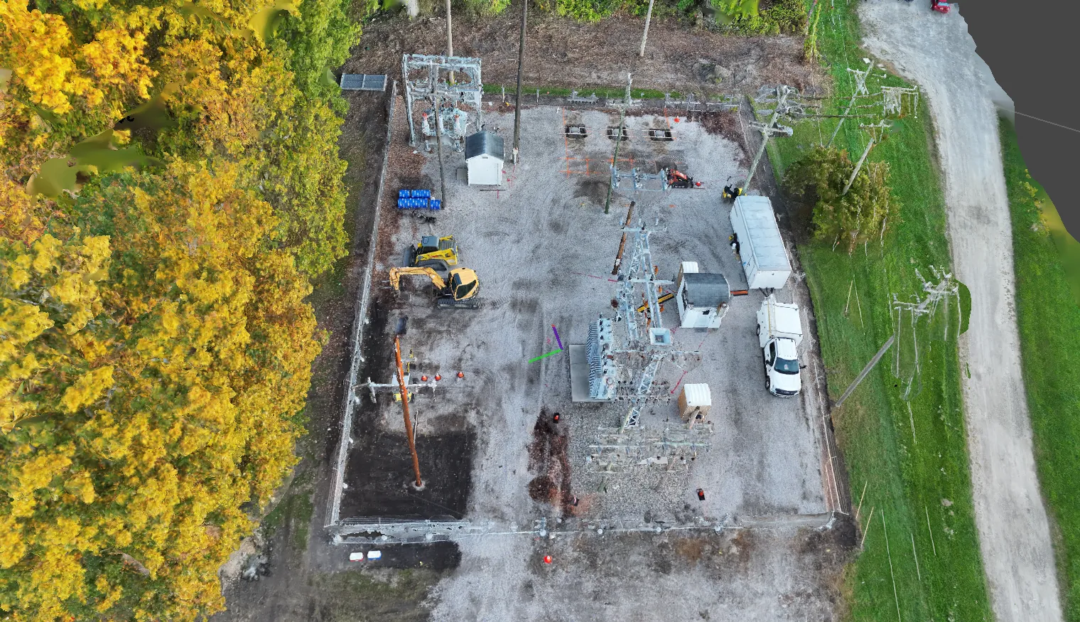 Top-down drone orthomosaic of a substation yard under construction, with an excavator, crew trucks and equipment inside the fence and fall-colored trees alongside